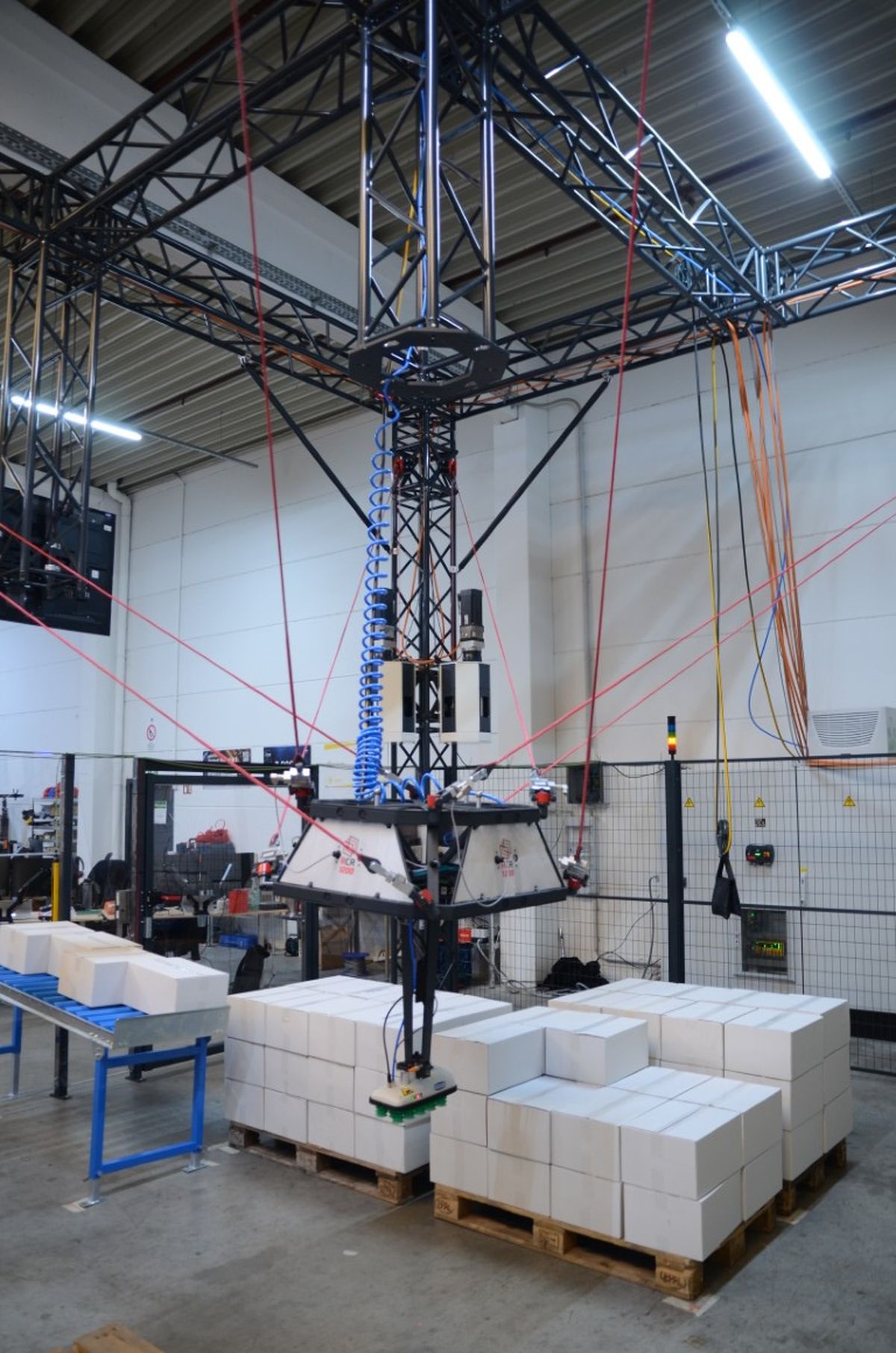 A cable-driven robot operates above pallets with white boxes in an industrial hall. Multiple tensioned cables control the gripper, enabling automated material handling and order picking.