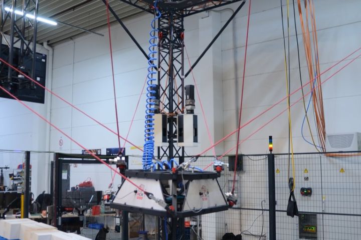 A cable-driven robot operates above pallets with white boxes in an industrial hall. Multiple tensioned cables control the gripper, enabling automated material handling and order picking.