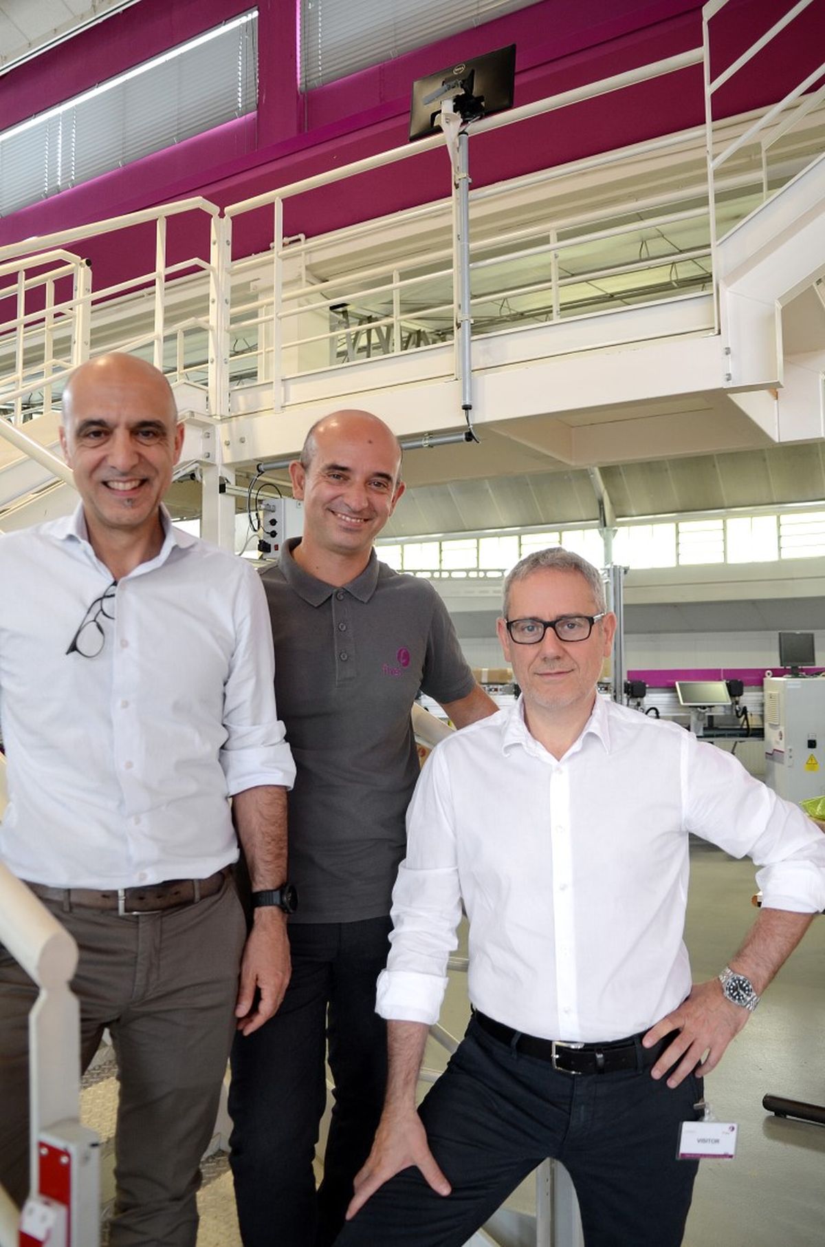 Tre persone in un ambiente industriale moderno sorridono alla fotocamera – simbolo di lavoro di squadra, collaborazione e competenza professionale in ambito industriale.