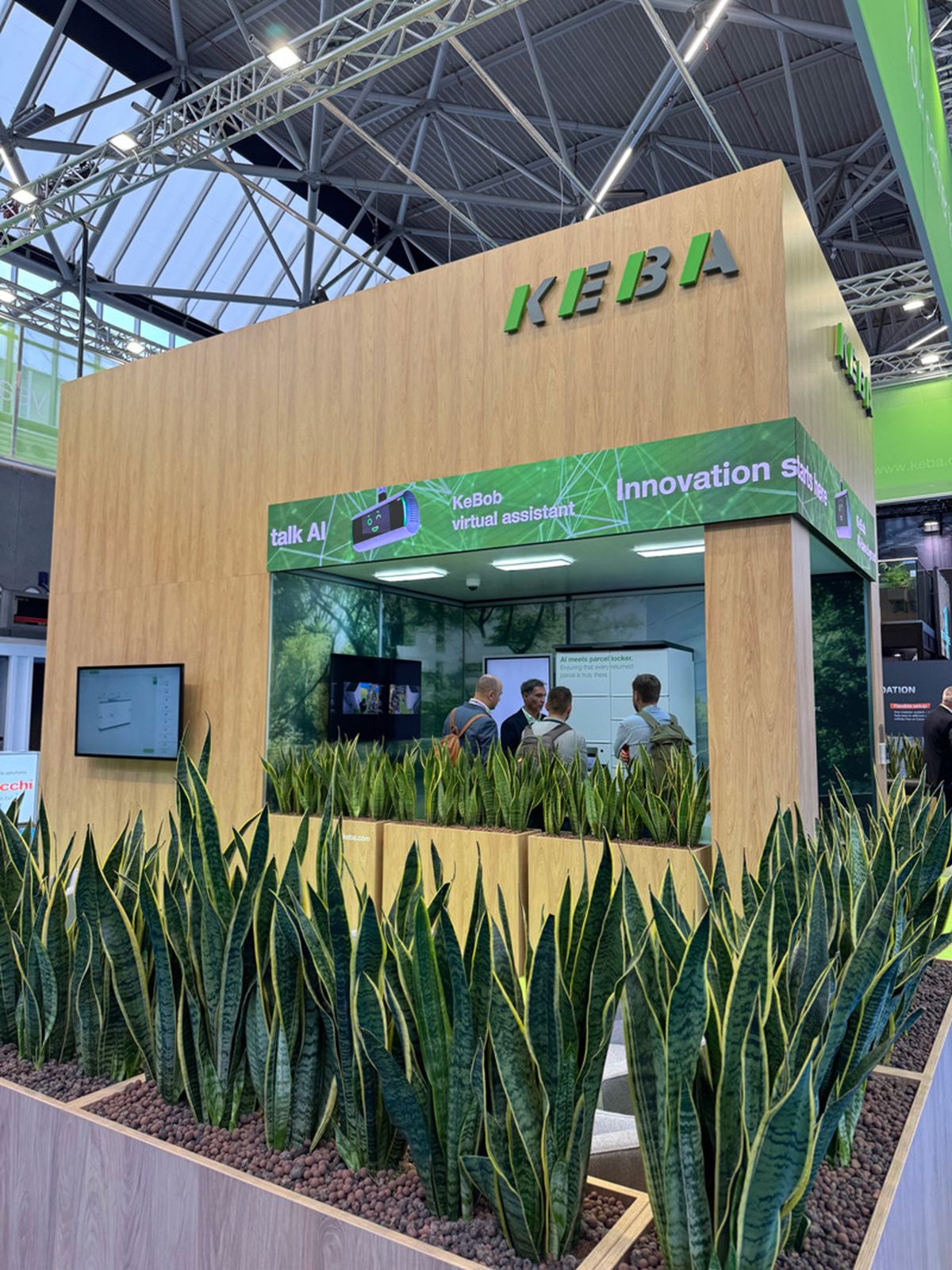 KEBA exhibition booth at Parcel + Post Expo 2025 with green plants, wooden walls, and a digital screen.
