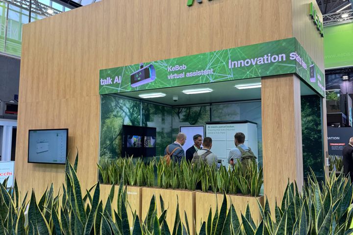 KEBA exhibition booth at Parcel + Post Expo 2025 with green plants, wooden walls, and a digital screen.