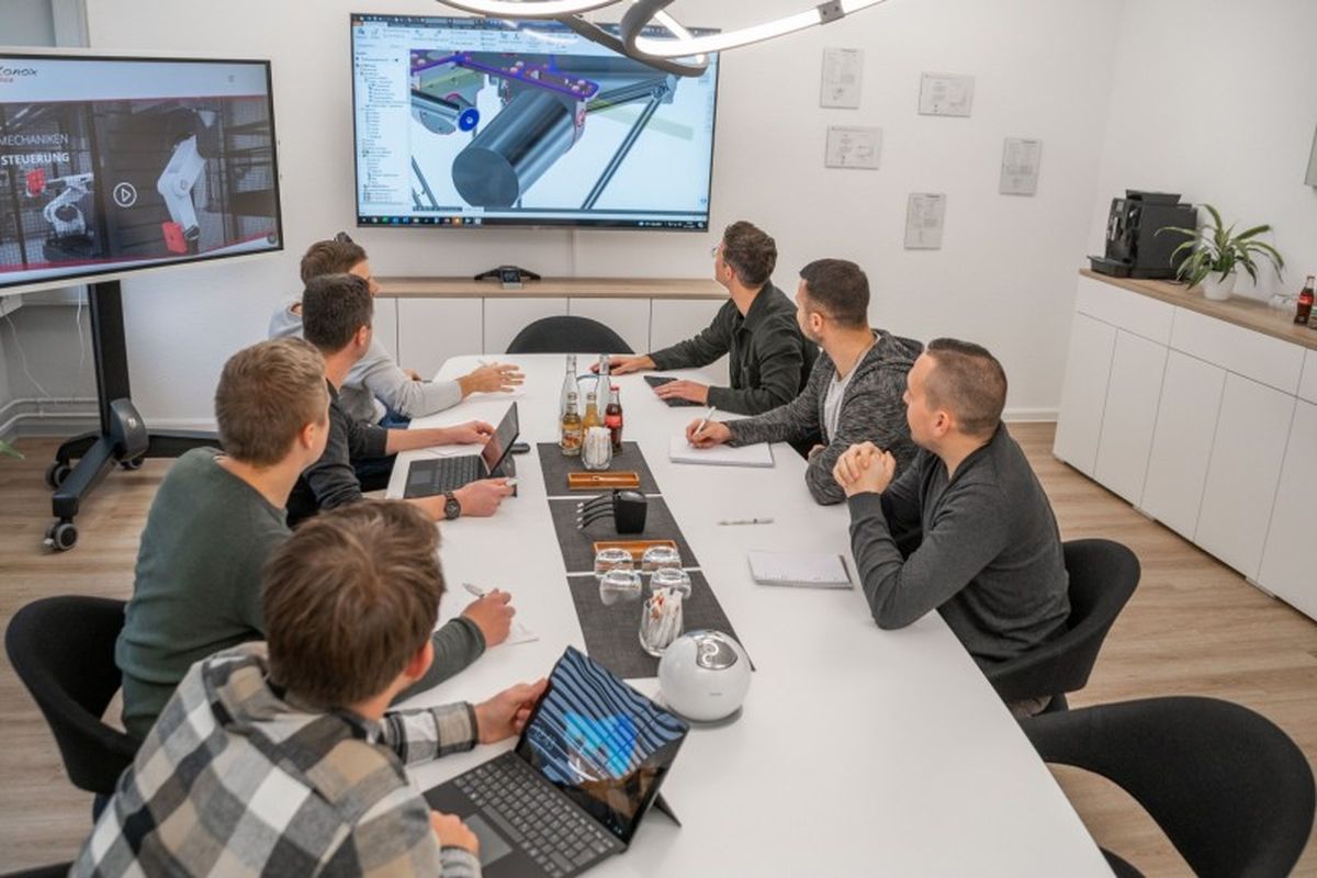 Ein autonox-Team hält eine Besprechung im Besprechungszimmer ab. Sie blicken auf den Screen an der Wand, auf dem ein grafische Detailzeichnung eines autonox Delta-Roboters zu sehen ist.