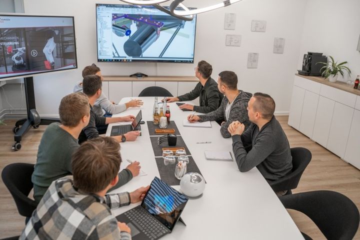 An autonox team is holding a meeting in the conference room. They are looking at the screen on the wall, which displays a detailed graphic drawing of an autonox Delta robot.
