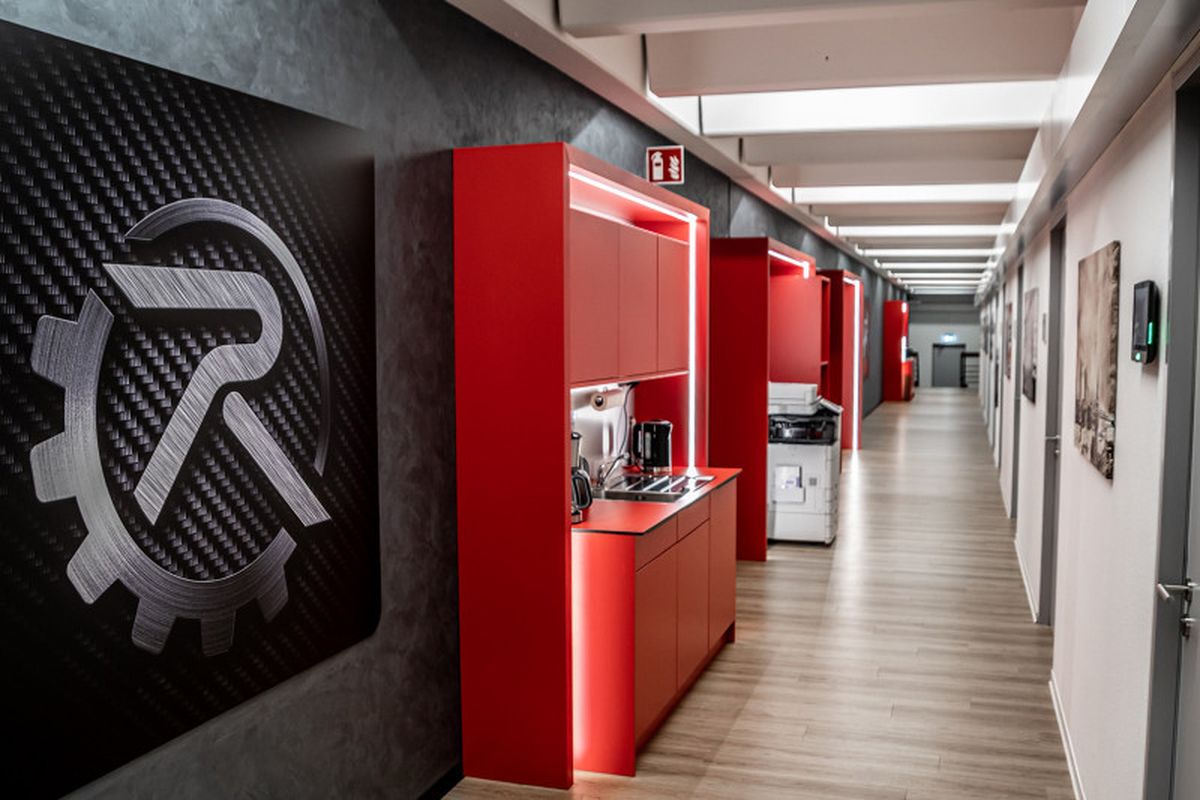 Ein Bild aus den Büroräumen von autonox. Zu sehen ist ein langer Gang, rechts sieht man graue Türen. Links im Vordergrund ist groß das Logo zu sehen, dann rote Küchenschränke und ein Kopierer. 