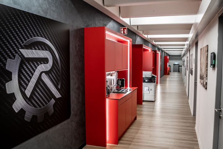 A picture from the autonox offices. It shows a long corridor with gray doors on the right. The logo is prominently displayed on the left in the foreground, followed by red kitchen cabinets and a copier.