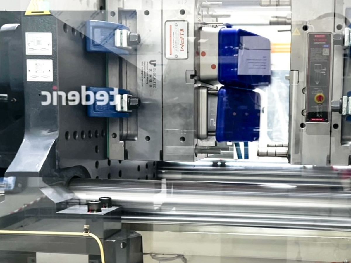 Close-up view through a transparent safety cover of a Tederic injection molding machine in action, with blue mold components and mechanical parts clearly visible.