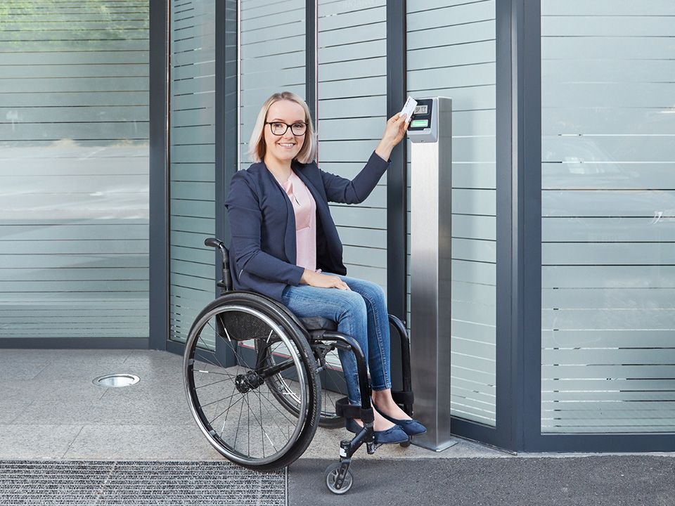 Eine Frau im Rollstuhl benutzt ihre Zugangskarte, um die automatische Drehtür eines modernen Bürogebäudes zu öffnen.