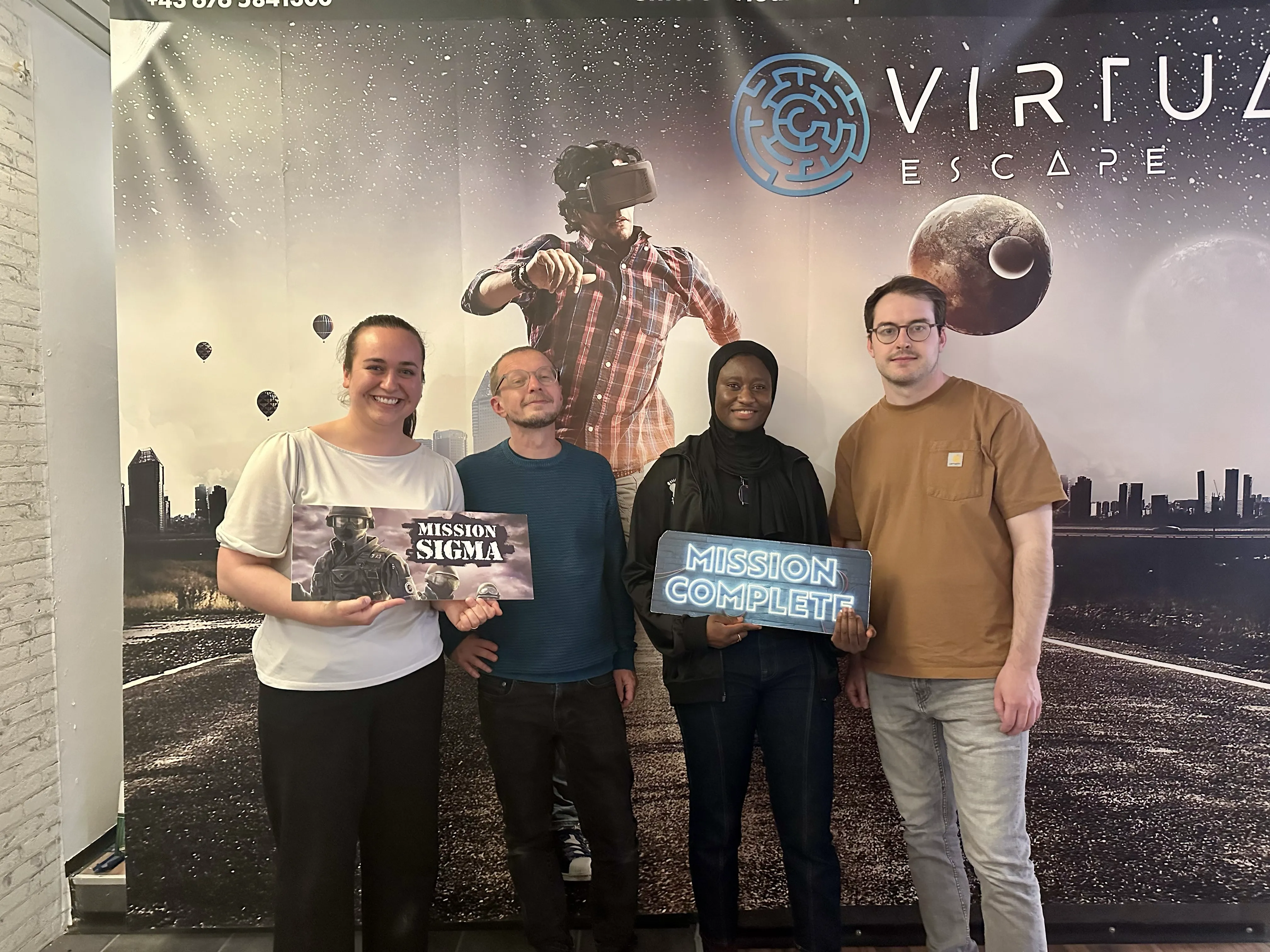 Four people stand in front of a "Virtual Escape" backdrop, holding "Mission Sigma" and "Mission Complete" signs, looking pleased.