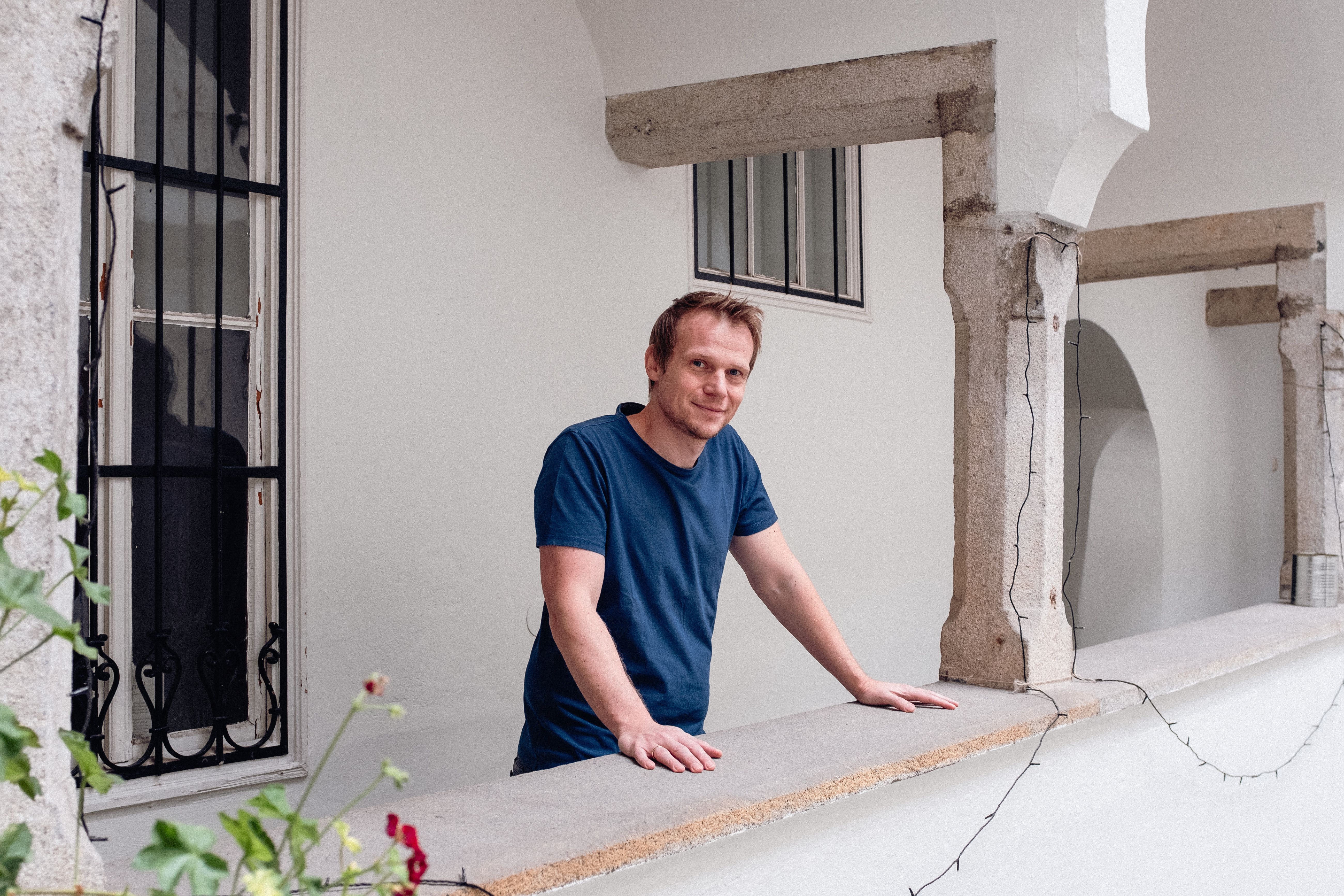 Helmut leans on a white balcony railing, with architectural columns and a window in the background (Hauptplatz 23 location).