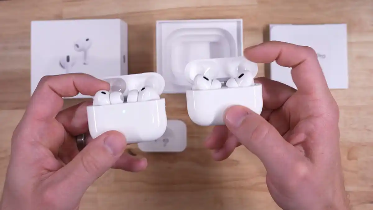 Bird's eye view of two hands, the left one holding the AirPods Pro 3 seated in their MagSafe charging case, the right one holding the AirPods Pro 2 seated in their MagSafe charging case, both with an open lid.