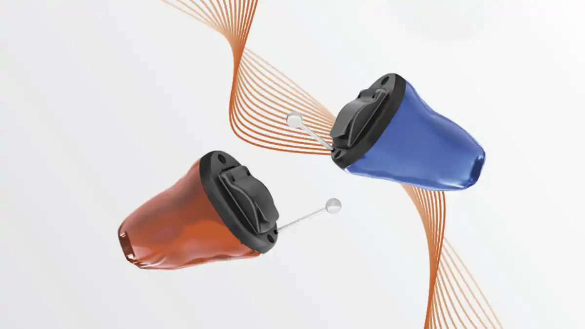 Two tiny in-ear hearing aids, one red and one blue, with an orange ribbon-like sound wave in background