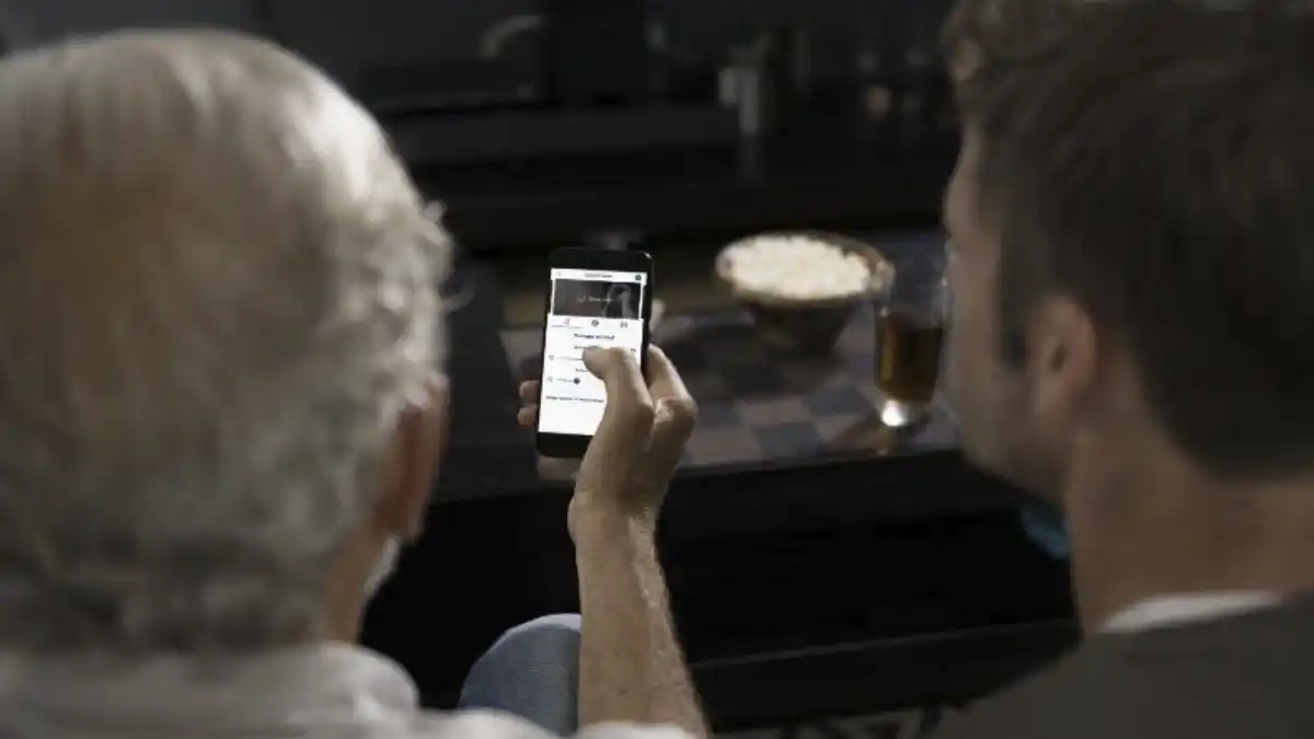Photo of man adjusting his hearing aids with Miracle-Ear app with popcorn and TV in background