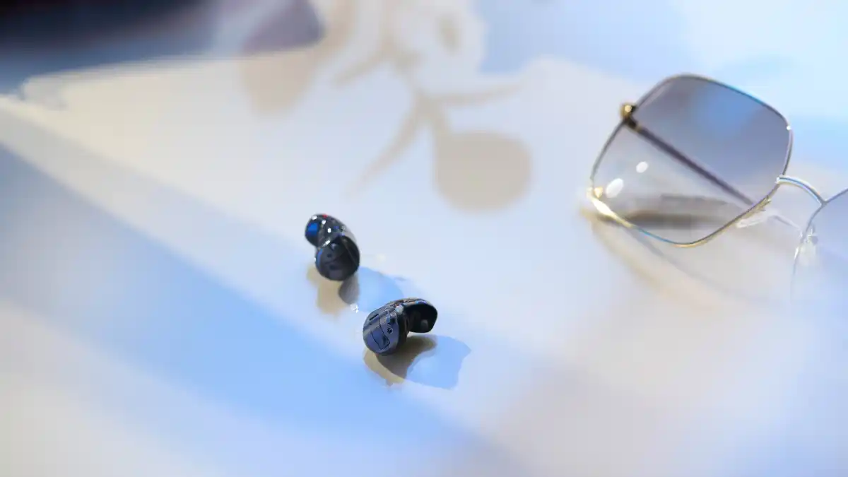 Two tiny black CIC hearing aids next to sunglasses.