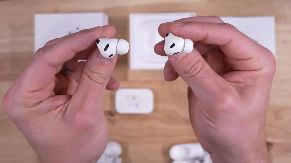 Bird's eye view of two hands, the left one holding an Apple AirPods Pro 3 earbud and the right one holding an AirPods Pro 2 earbud with the box and accessories sitting on the table in the background underneath.