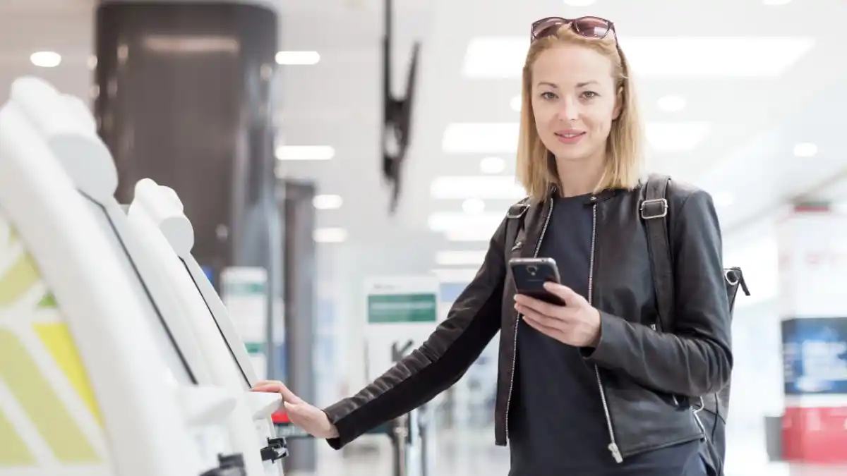Woman holding a phone standing next to an airport kiosk.