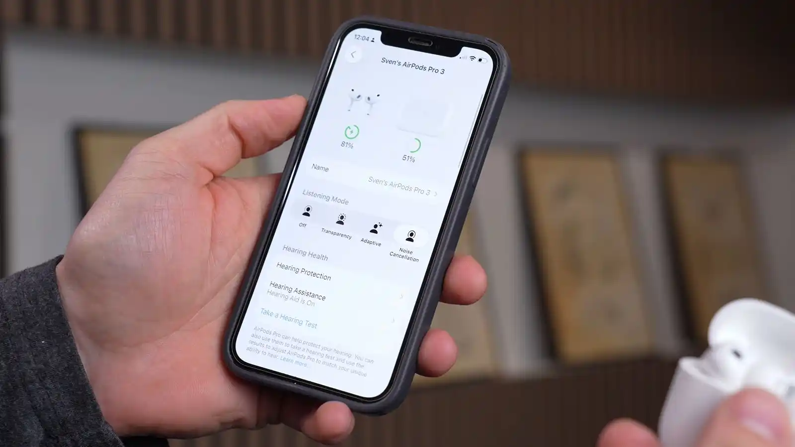 Left hand holding an iPhone and navigating the AirPods hearing aid features, while the right hand holds an open AirPods MagSafe charging case with AirPods Pro 3 earbuds inside.