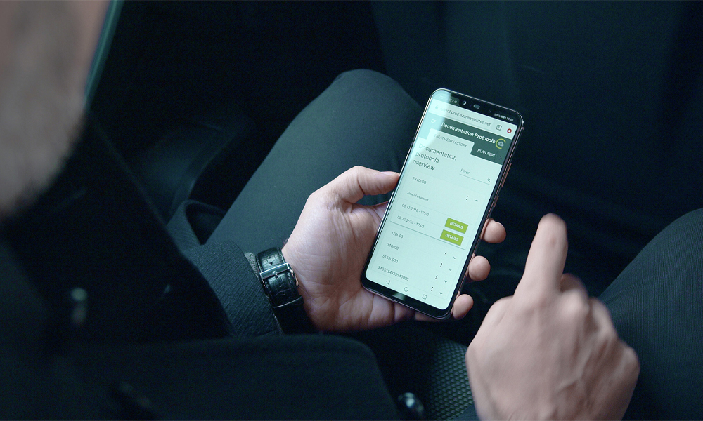 Una persona sostiene un teléfono inteligente, en la pantalla se puede ver la plataforma IoDent®, con un fondo sombreado.