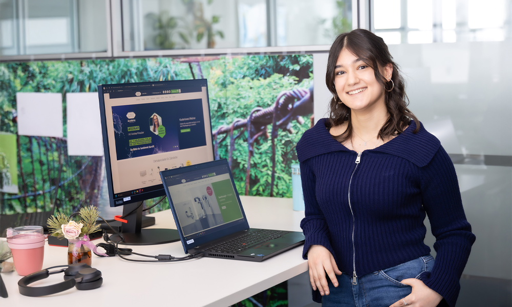 Zeynep Tosun lächelt vor ihrem Büroarbeitsplatz und ihrem Computer in die Kamera.