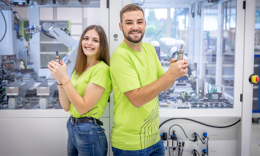 Eine Zerspanungstechnikerin und ein Zerspanungstechniker vor einer CNC-Maschine, halten Bohrer und lächeln in die Kamera