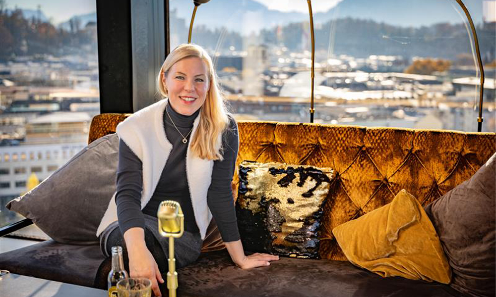 PD Dr. Amely Hartmann lächelt in die Kamera, sitzt auf einer Bank, im Hintergrund ist durch ein Fenster die Stadt Salzburg zu sehen.
