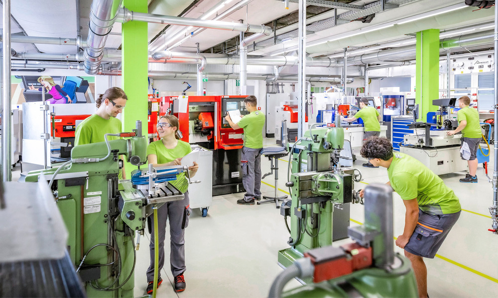 Der W&H Campus mit Lehrlingen in grüner Arbeitskleidung, die an verschiedenen Werkzeugmaschinen arbeiten und sich austauschen.