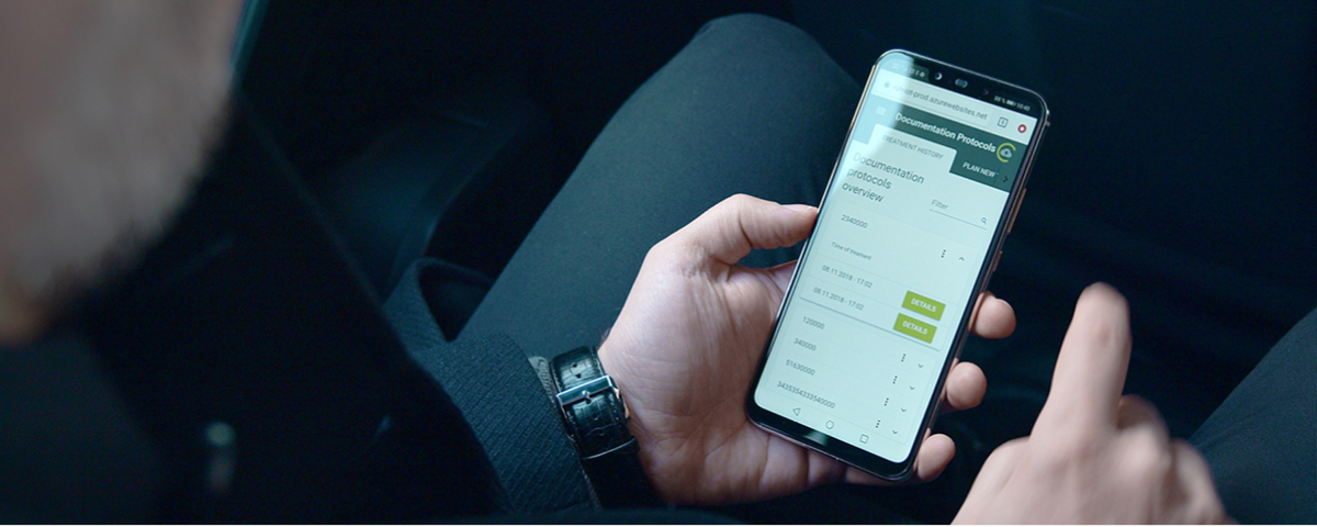 Una persona tiene in mano uno smartphone che mostra l'interfaccia dei protocolli di documentazione W&H ioDent®, con i registri di sterilizzazione e i dettagli della documentazione del dispositivo.