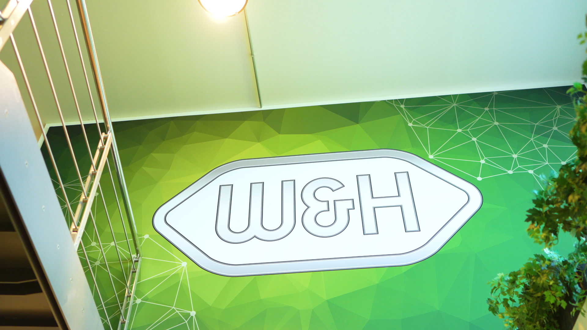 Logo W&H su una parete interna verde con elementi grafici geometrici e struttura di una scala metallica sul lato sinistro.