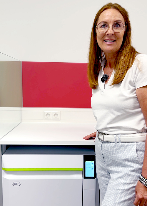 Andrea Knauber steht in einer modernen Zahnarztpraxis neben dem Thed-Thermodesinfektor von W&H. Im Hintergrund sind ein heller Arbeitsbereich und eine farbige Wandgestaltung zu sehen. Andrea Knauber blickt in die Kamera und lächelt freundlich.