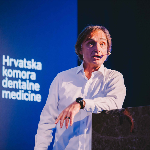 Speaker Istvan Urban presenting on stage at the PERIOIMPLANT10 event, wearing a white shirt and headset microphone, standing in front of a blue conference backdrop.