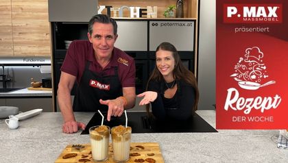 Philipp und Tanja präsentieren in einer modernen Küche zwei Gläser Dalgona-Kaffee mit der charakteristischen cremigen Schaumkrone und Strohhalmen. Vor ihnen auf einem Holzbrett liegen die fertig zubereiteten Dalgona-Zuckerplätzchen zusammen mit einer Stern-Ausstechform.