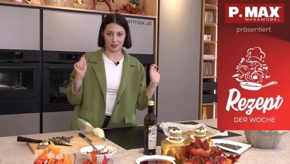 Die Köchin Sarah lächelt in die Kamera, während sie in einer modernen Küche kleine Kürbis-Törtchen mit einer hellen Creme aus einem Spritzbeutel verziert. Auf der Arbeitsfläche stehen neben den fertigen Törtchen eine Flasche steirisches Kürbiskernöl und herbstliche Dekoration.