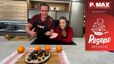 Tanja und Philipp präsentieren lächelnd den fertigen „No bake - Black Cheesecake“ in einer modernen P.MAX Küche. Der mit dunklen Keksstücken und Orangenabrieb verzierte Kuchen steht im Vordergrund auf einem Holzbrett, umgeben von frischen Orangen.