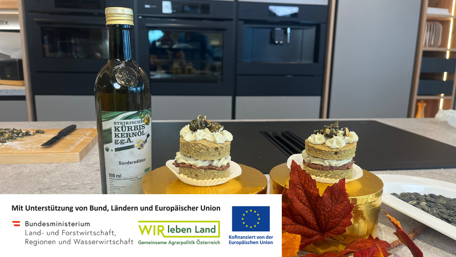 Ein fertig angerichtetes Kürbis-Törtchen, das aus zwei Schichten hellem Kuchenboden besteht. Dazwischen und obendrauf ist eine grünliche Kürbiskernöl-Creme aufgespritzt und das Törtchen ist mit gehacktem Kürbiskern-Krokant garniert.