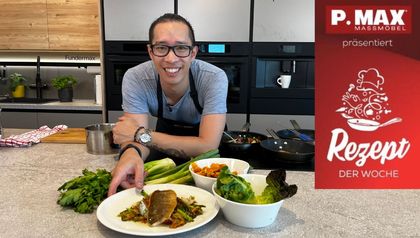 Koch Peter Lehner präsentiert in einer modernen Küche das fertige Gericht: gebratener Saibling auf einem Bett aus Gemüse. Auf der Arbeitsplatte stehen weitere Zutaten wie Romanasalat und gebratene Pilze bereit.