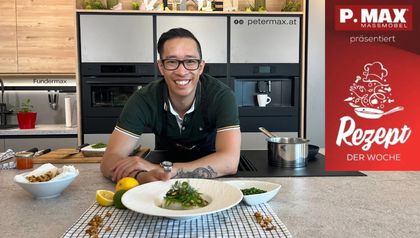 Koch Peter Lehner präsentiert lächelnd in einer modernen Küche den fertig angerichteten Kabeljau mit Riesling Beurre Blanc. Das Gericht ist auf einem weißen Teller angerichtet und mit Zuckerschotensalat sowie Croutons garniert, umgeben von weiteren frischen Zutaten auf der Kücheninsel.