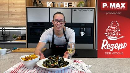 Chefkoch Peter Lehner präsentiert lächelnd die fertig zubereiteten Moules Frites in einer modernen P.MAX Küche. Vor ihm auf der Arbeitsplatte sind ein Teller mit Miesmuscheln in Sud, eine Schüssel goldbraune Pommes Frites und ein Glas Weißwein auf einem rot-weiß karierten Tuch angerichtet.