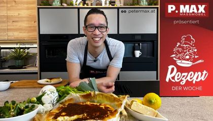 Peter Lehner lächelt in einer modernen Küche hinter einer frisch gebackenen Spinat-Quiche, die zusammen mit Zutaten wie Lauch, Zitrone und Käse auf der Arbeitsplatte angerichtet ist.