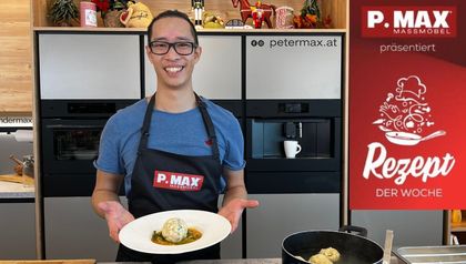 Spitzenkoch Peter Lehner präsentiert lächelnd einen Teller mit dem fertigen Kalbsrahmgulasch und einem Semmelknödel in einer modernen Küche. Er trägt eine schwarze Schürze und im Vordergrund ist ein Topf mit kochendem Wasser und weiteren Knödeln zu sehen.
