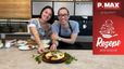 Koch Peter Lehner und Sarah Jahn präsentieren lächelnd die fertige Pizza mit Pistazienpesto in einer modernen Küche. Im Vordergrund liegt die Pizza, belegt mit Pesto, marinierten Tomaten und Mozzarella, auf einem Holzbrett.