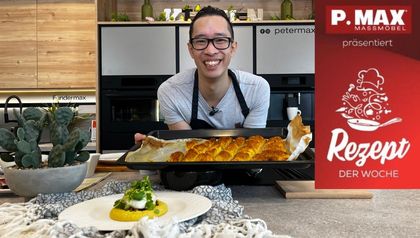 Chefkoch Peter Lehner steht lächelnd in einer modernen Küche und präsentiert ein Backblech mit goldbraunem Gebäck. Im Vordergrund steht ein Teller mit dem fertig angerichteten Gericht Hamshuka, bestehend aus Hummus, würzigem Hackfleisch und einem pochierten Ei, das mit frischen Kräutern garniert ist.