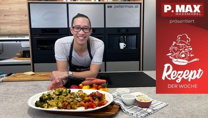 Koch Peter Lehner präsentiert lächelnd eine Platte mit den drei vegetarischen Ofengemüse-Gerichten: Brokkoli mit Mandeln, Karotten mit Sesam und Kohlsprossen mit Erdnüssen. Er steht in einer modernen Küche vor hochwertigen Einbaugeräten, wobei die verschiedenen Zutaten dekorativ auf der Arbeitsplatte angerichtet sind.