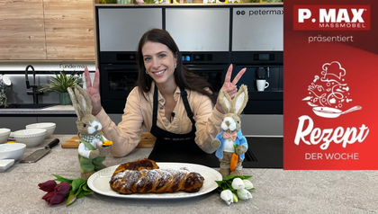 Tanja Pfaffeneder präsentiert lächelnd einen frisch gebackenen, goldbraunen Osterzopf in Hasenform, der mit Puderzucker bestäubt auf einer weißen Servierplatte liegt. Die Szene in der modernen Küche ist passend zum Rezept mit dekorativen Osterhasen-Figuren und bunten Tulpen gestaltet.