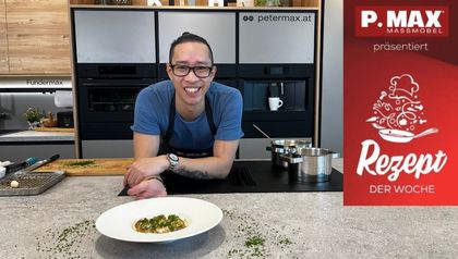 Peter Lehner präsentiert die fertig angerichteten Jakobsmuscheln mit Sauce auf einem weißen Teller, garniert mit fein gehackten Kräutern. Er steht lächelnd in einer modernen Küche hinter einer Arbeitsplatte, auf der im Hintergrund Töpfe auf dem Herd und Vorbereitungsutensilien zu sehen sind.