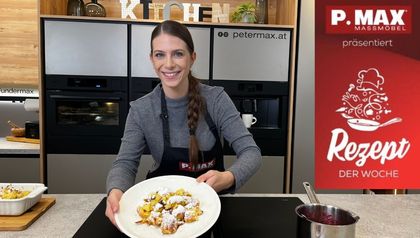 Tanja präsentiert einen Teller mit frisch zubereitetem, mit Staubzucker bestreutem Kaiserschmarrn. Sie steht in einer modernen Küche mit dunklen Einbaugeräten und einem dekorativen „KITCHEN“-Schriftzug im Hintergrund.