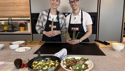 Sängerin Virginia Ernst und Peter Lehner stehen lächelnd in einer modernen Küche und halten frischen grünen Spargel in den Händen. Im Vordergrund sind die fertig zubereiteten Gerichte auf einem Holzbrett angerichtet: eine Spargel-Frittata direkt in der Pfanne sowie ein bunter Spargelsalat mit in Schinken gewickelten Stangen in einer weißen Schüssel.