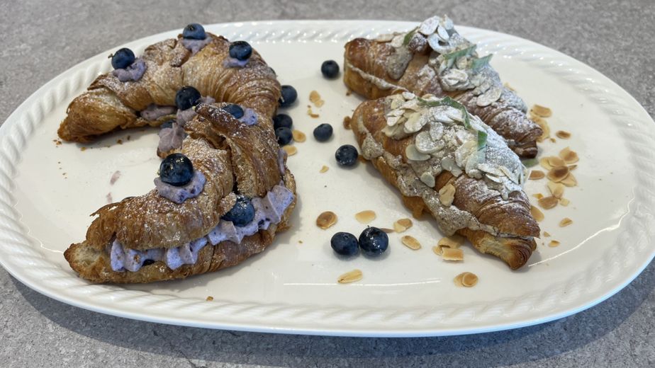 Die fertigen Mandel- und Heidelbeer-Croissants aus dem Rezept, angerichtet auf einer weißen Platte. Die Heidelbeer-Croissants sind mit einer lila Creme und frischen Beeren gefüllt, die Mandel-Croissants sind mit Mandelblättchen bestreut.
