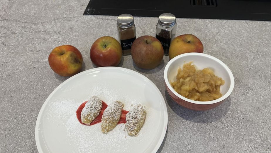 Drei mit Staubzucker bestreute Topfennockerln auf einem roten Fruchtspiegel, serviert neben einer Schüssel mit stückigem Apfelmus. Im Hintergrund ergänzen vier frische Äpfel und Gewürzdosen wie Zimt das fertig angerichtete Dessert auf einer grauen Küchenarbeitsplatte.