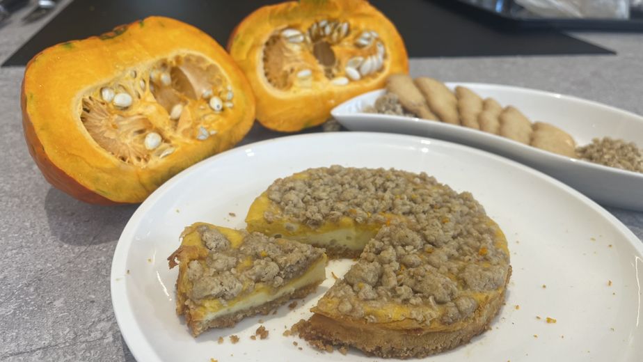 Ein fertiger Kürbis-Cheesecake mit Streuseln auf einem weißen Teller. Ein Stück ist bereits herausgeschnitten und zeigt die helle und orangefarbene Füllung. Im Hintergrund sind ein aufgeschnittener Kürbis und Spekulatius-Kekse als Zutaten zu sehen.