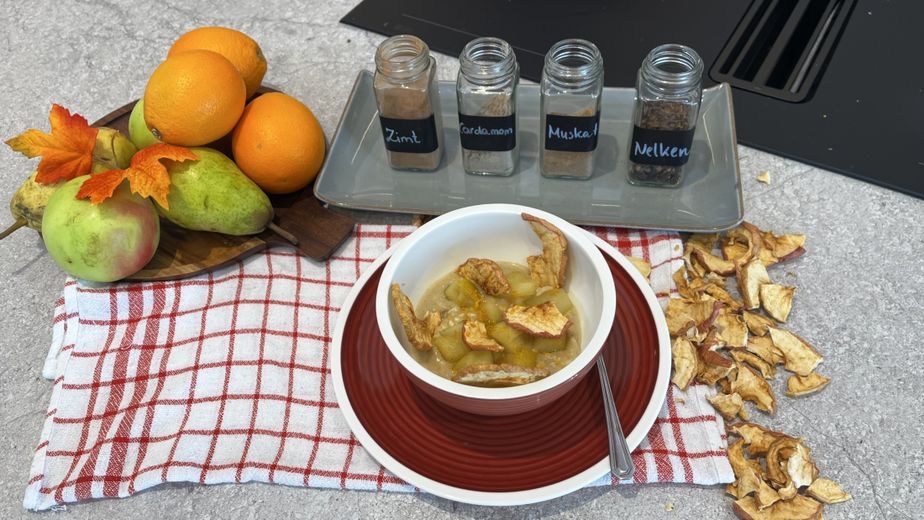 Eine Schüssel mit veganem Milchreis und Birnenkompott, garniert mit getrockneten Apfelchips. Im Hintergrund sind die Zutaten für das herbstliche Gericht arrangiert: frische Äpfel, Birnen und Orangen sowie Gewürzgläser mit Zimt, Kardamom, Muskat und Nelken.