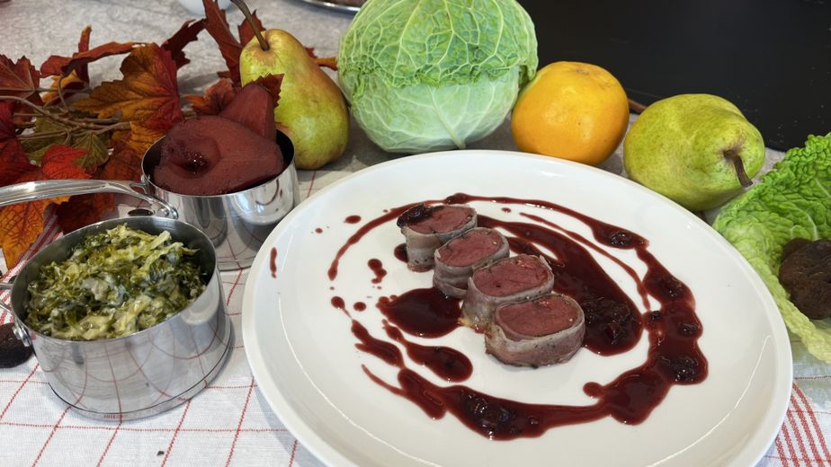 Vier Medaillons vom Rehrücken im Speckmantel, angerichtet auf einem weißen Teller und mit einer dunklen Soße beträufelt. Daneben stehen in kleinen Edelstahltöpfen der fertige Rahmwirsing und eine pochierte Rotweinbirne.